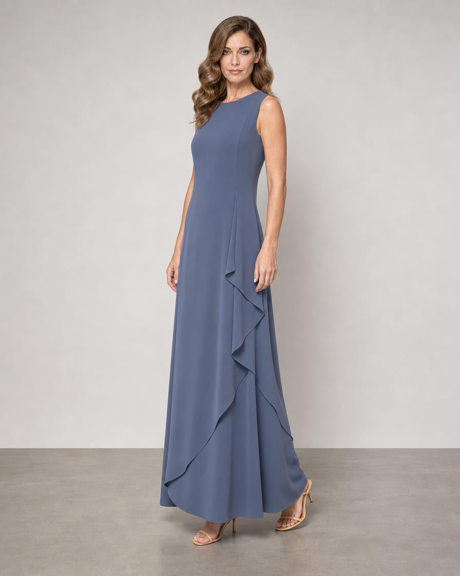 A woman modeling a sleeveless, floor-length blue-gray gown with a ruffled asymmetrical hem, standing against a neutral studio backdrop.