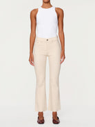A model wearing a pair of high-waisted, light beige cropped flare jeans with a white sleeveless top and dark brown pointed flats, shot against a solid off-white background.
