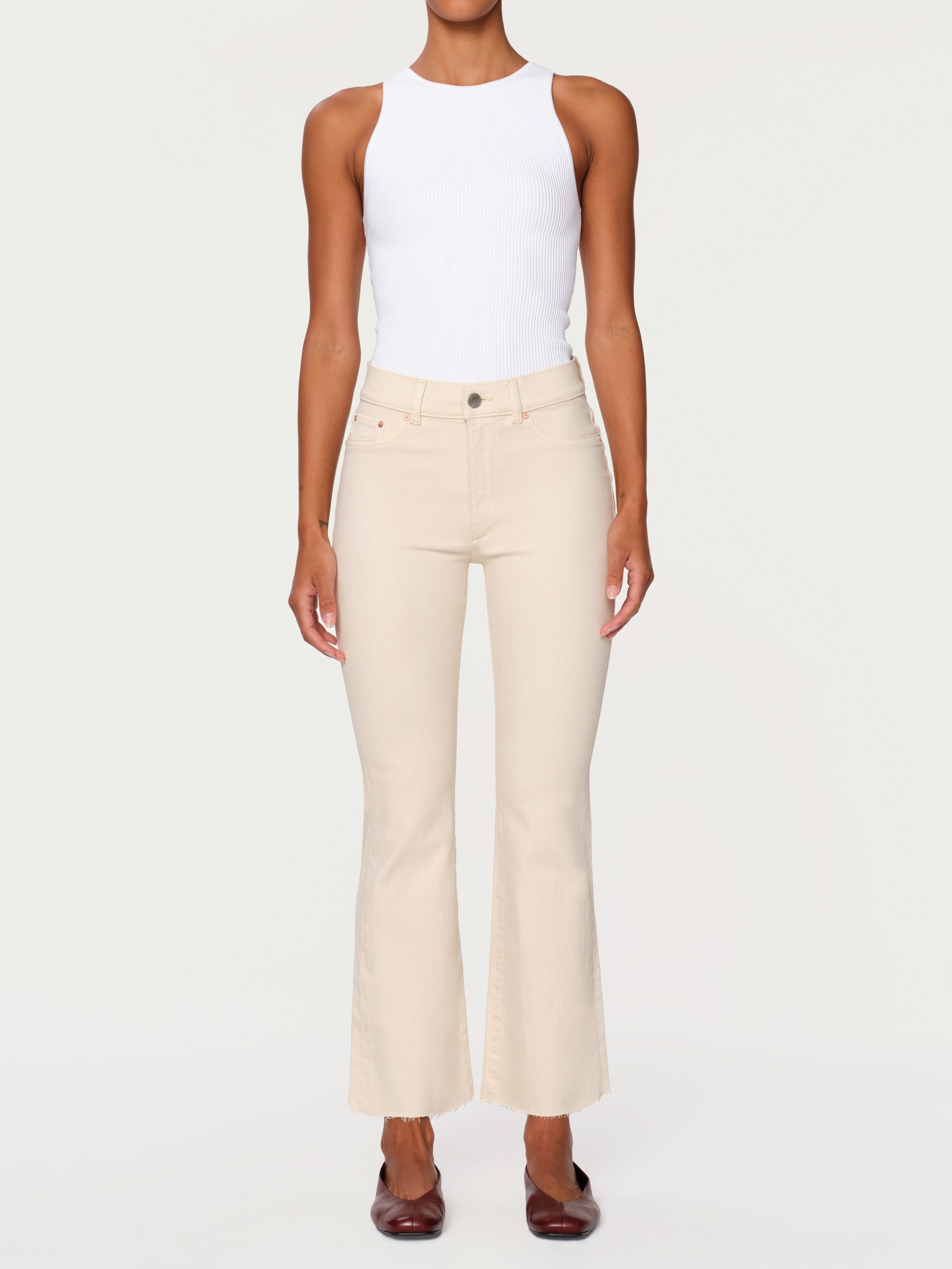 A model wearing a pair of high-waisted, light beige cropped flare jeans with a white sleeveless top and dark brown pointed flats, shot against a solid off-white background.