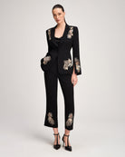 A model wears a black two-piece suit featuring ornate gold and beige floral embroidery on the blazer's shoulders, sleeves, and pockets, paired with matching cropped trousers and black high heels, set against a plain light gray studio background.