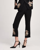 Black cropped trousers with ornate floral embroidery on the pockets and lower hems, shown on a model wearing black pointed T-strap heels against a light gray studio background.