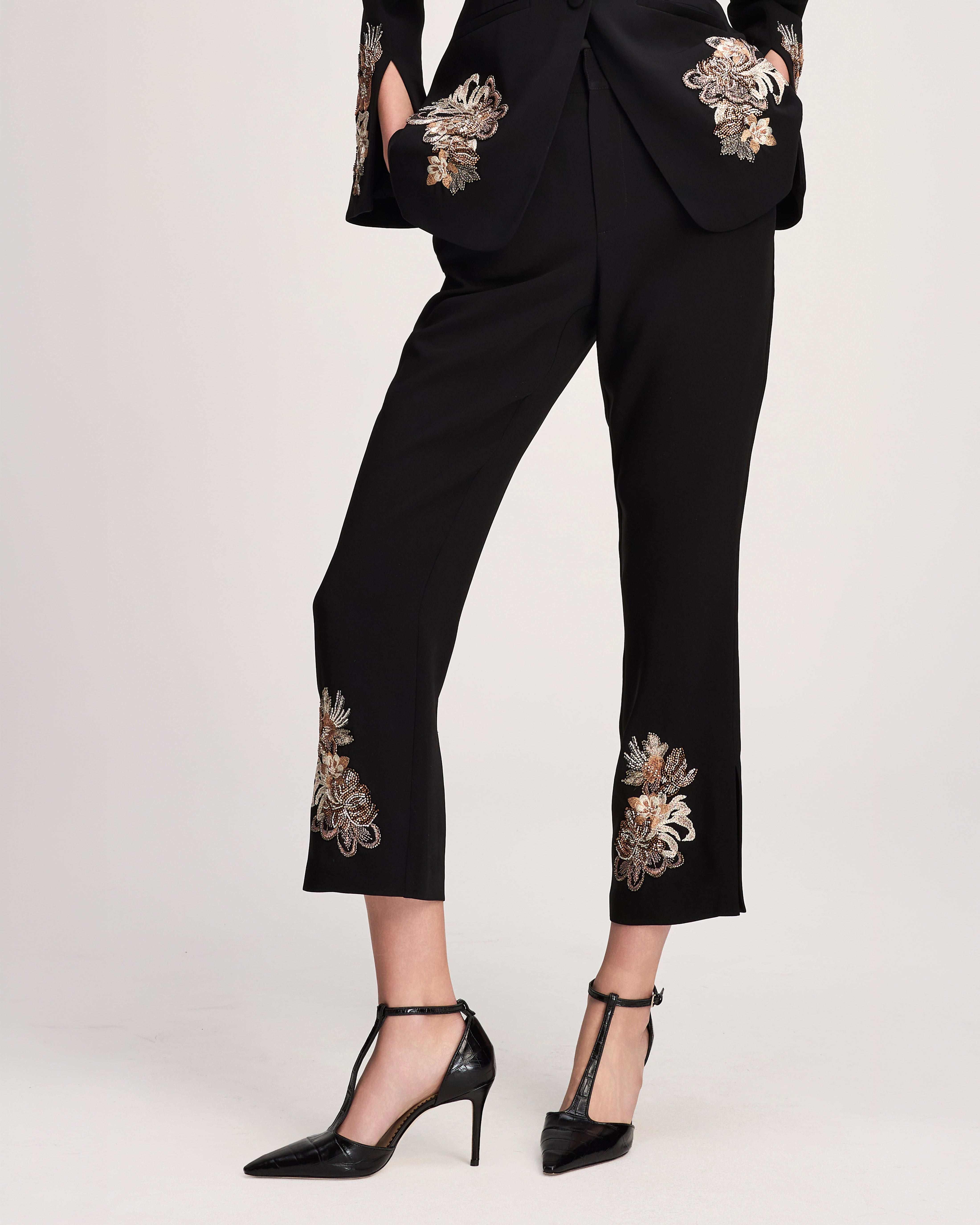 Black cropped trousers with ornate floral embroidery on the pockets and lower hems, shown on a model wearing black pointed T-strap heels against a light gray studio background.