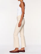 A side-profile view of a model wearing cream-colored flared pants, a white tank top, and dark brown leather flats against a plain white studio background.