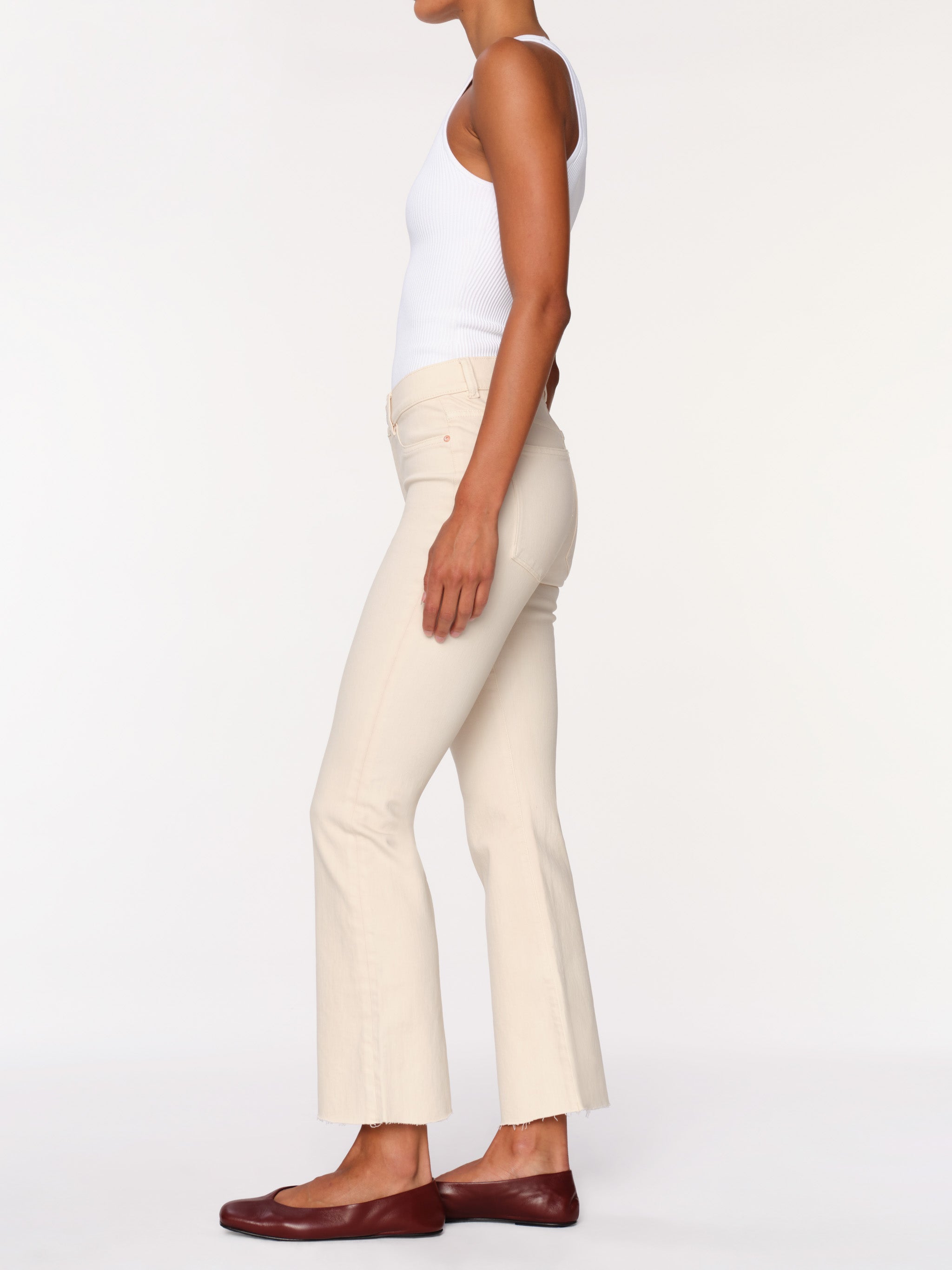 A side-profile view of a model wearing cream-colored flared pants, a white tank top, and dark brown leather flats against a plain white studio background.