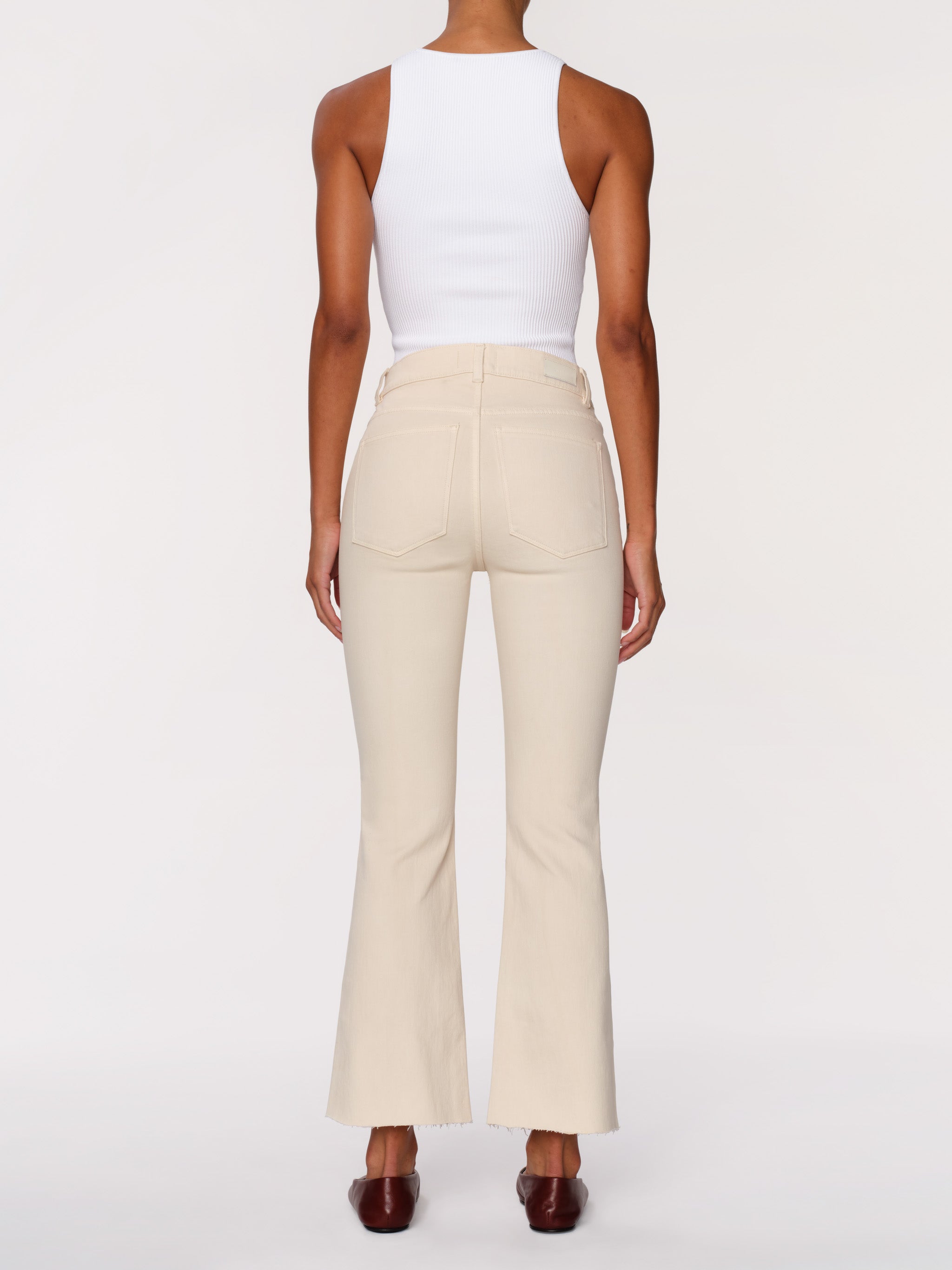 A model shown from the back wearing high-waisted cream-colored flared pants and a white sleeveless top, paired with dark brown shoes, against a plain light gray background.
