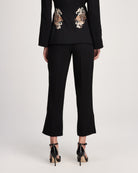 A model shown from the back wearing a black cropped blazer with ornate gold and white floral embroidery on the upper back, paired with black cropped trousers and black patent high heels, against a light gray studio background.