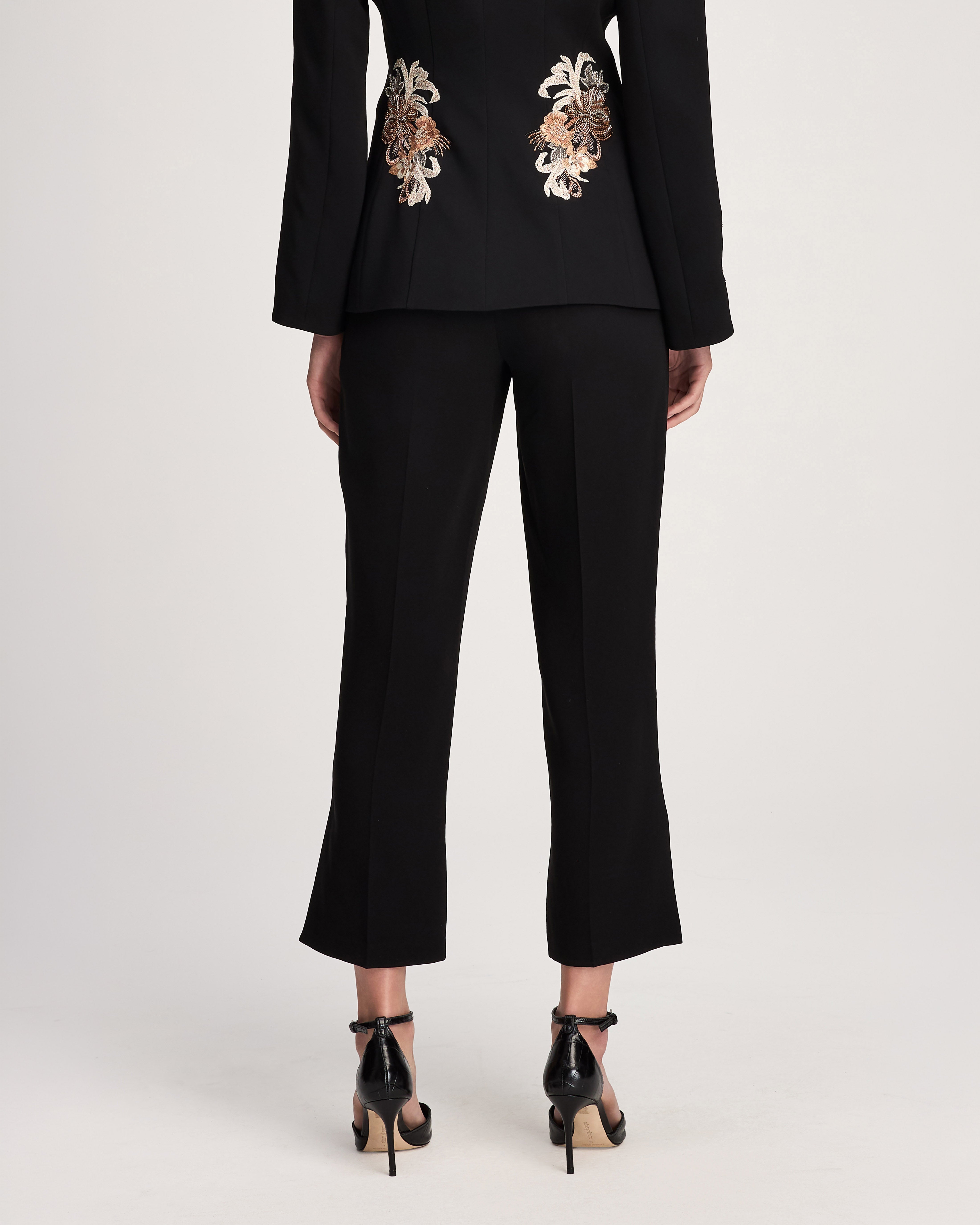 A model shown from the back wearing a black cropped blazer with ornate gold and white floral embroidery on the upper back, paired with black cropped trousers and black patent high heels, against a light gray studio background.