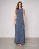 A woman modeling a sleeveless, floor-length blue-gray dress with a round neckline and layered ruffle detail along the side, standing against a neutral studio backdrop.