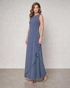 A woman modeling a sleeveless, floor-length blue-gray gown with a ruffled asymmetrical hem, standing against a neutral studio backdrop.