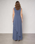A woman viewed from the back wearing a sleeveless, floor-length blue dress with a draped asymmetrical hem, standing against a plain light gray studio wall.