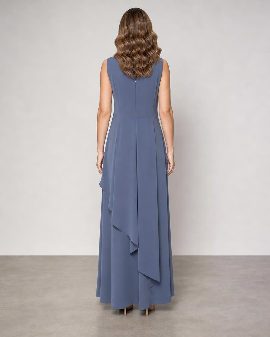 A woman viewed from the back wearing a sleeveless, floor-length blue dress with a draped asymmetrical hem, standing against a plain light gray studio wall.