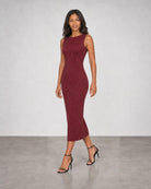 A woman modeling a sleeveless, bodycon midi dress in a textured burgundy fabric, paired with black strappy high-heeled sandals, set against a neutral gray studio background.