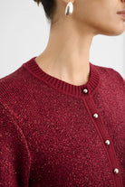 Close-up of a person wearing a textured burgundy knit cardigan sweater with silver-tone buttons, paired with a silver hoop earring against a light gray background.