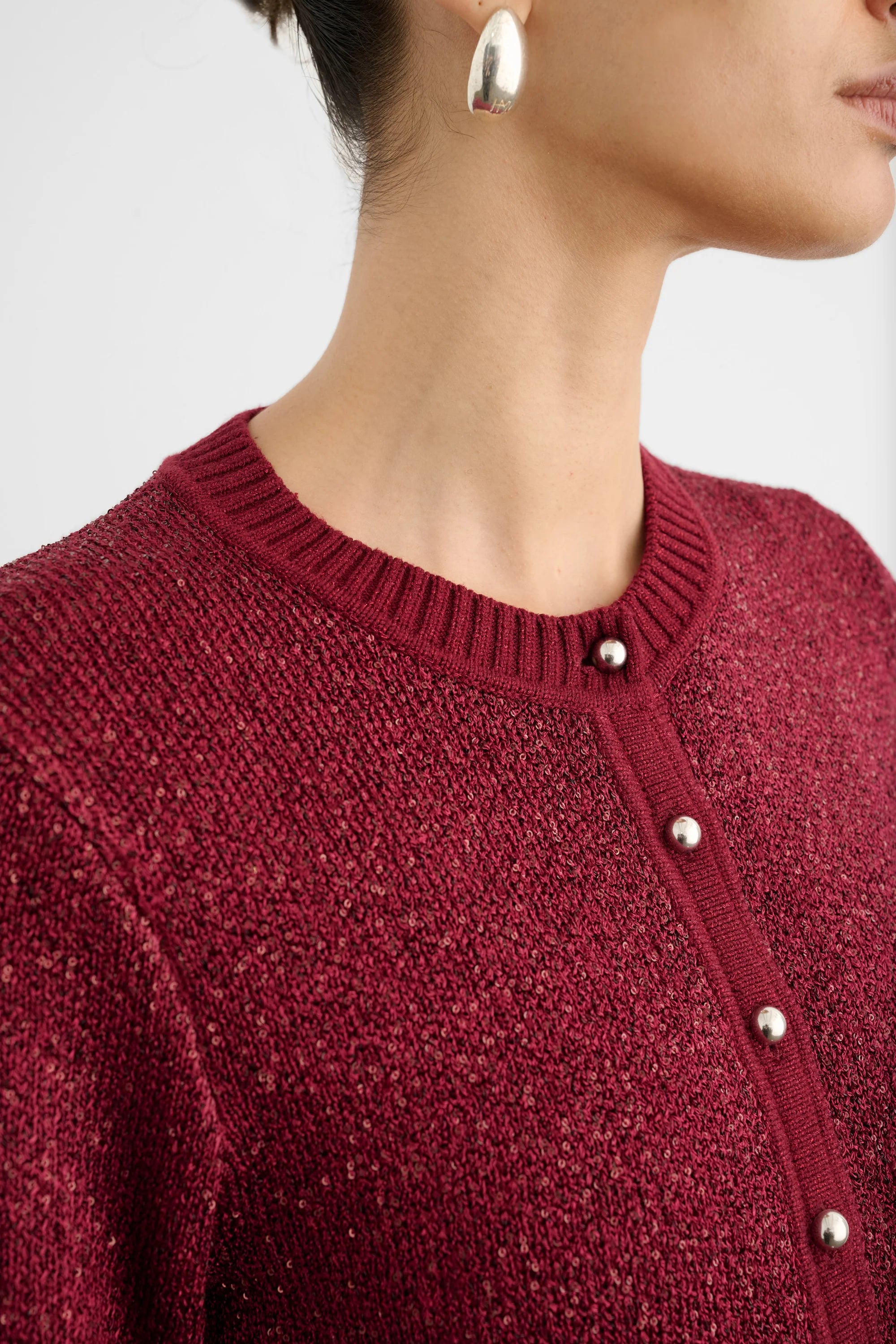 Close-up of a person wearing a textured burgundy knit cardigan sweater with silver-tone buttons, paired with a silver hoop earring against a light gray background.
