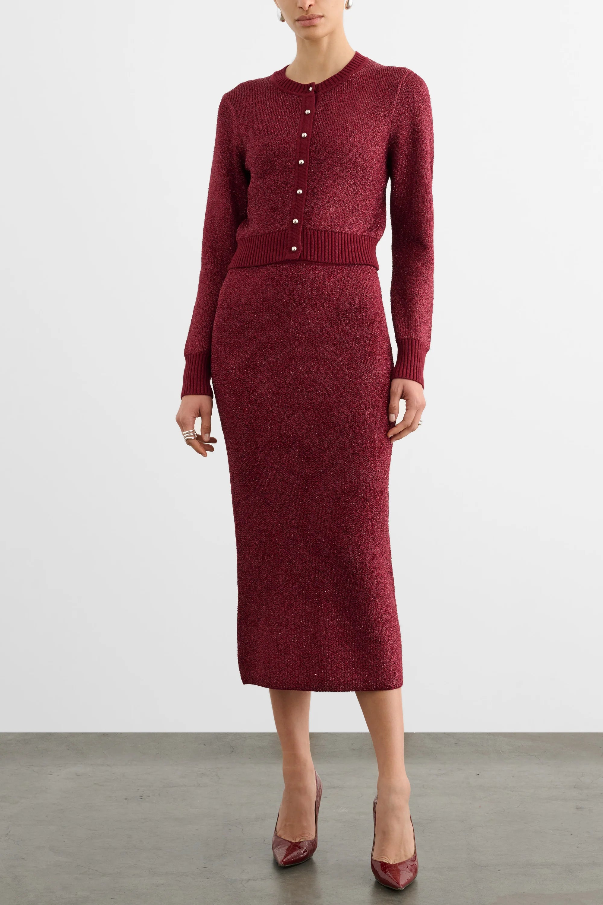 A model wearing a burgundy knit cardigan and matching midi skirt set, paired with matching burgundy heels, against a plain light gray studio background.