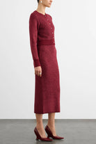 A model wearing a textured knit cardigan and matching midi skirt set in a deep burgundy red, paired with matching pointed-toe heels, shown from the side against a light gray studio background.