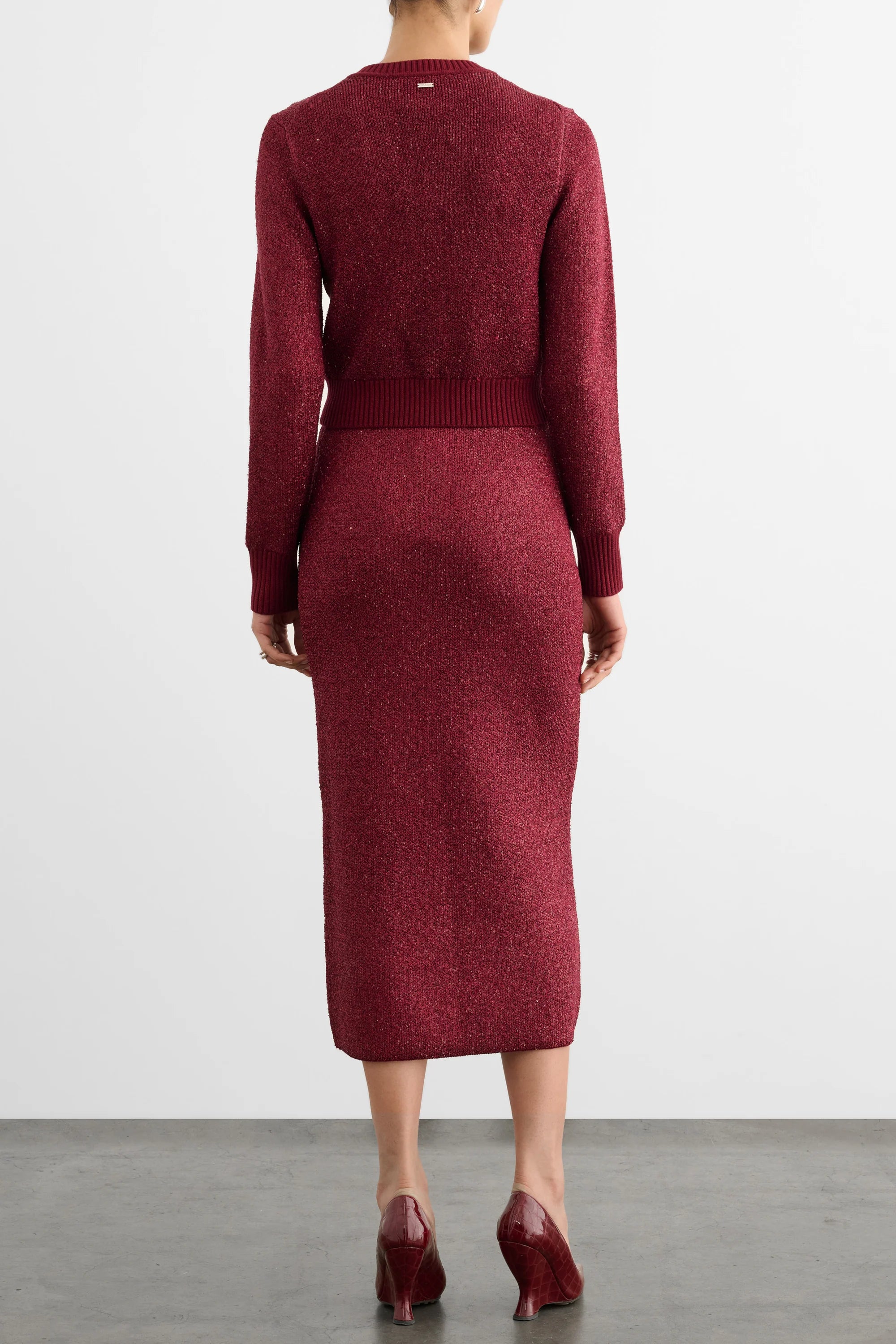 A model viewed from the back wearing a long-sleeve, crew-neck knit midi dress in a heathered burgundy red color, paired with matching burgundy high heels against a light gray studio background.